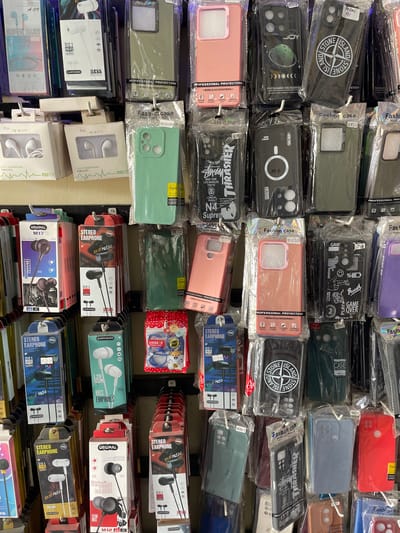 The image shows a display of mobile phone accessories in a shop, likely in Pasar Paloh, Indonesia. The display features rows of phone cases and earphones packaged in clear plastic and cardboard boxes. The phone cases are varied in color and design, with some featuring branded logos and graphics. The earphones are also packaged with brand names and specifications like "D21 Hi-Fi Audio" and "Bass Earphones". The overall environment suggests a retail setting where these items are being sold, with the products hung from hooks on a wall. The lighting is bright and evenly distributed, illuminating the colorful packaging and diverse range of products.