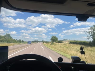 The image captures a driver's perspective from inside a truck on a highway in Rolampont, France. The scene is a sunny day with a bright blue sky dotted with fluffy white clouds. The highway stretches ahead, with a line of trucks visible in the distance. The right side of the road is lined with tall, dry grass and trees, suggesting a rural or semi-rural setting.

On the dashboard, a GPS device is mounted to the left, and what appears to be a dashcam is visible on the right, plugged into a power source. The steering wheel is in the foreground, obscuring part of the dashboard. The overall impression is one of travel and a journey in progress on a pleasant day. The time of day is clearly daytime, and the weather is fair. No people are directly visible in the frame, but the presence of the steering wheel and dashboard implies a driver. There is no discernible text in the image.