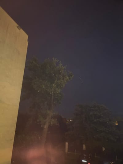 A nighttime exterior shot shows a portion of a light-colored building on the left side of the frame. The building is angled, with its facade taking up the left third of the image. The remaining two-thirds of the image are dominated by a dark, clear night sky. In the middle ground, several trees are visible, their foliage dark against the night sky. One tree in the center is taller and more defined, while others are shorter and more clustered. In the distance, faint lights suggest the presence of buildings or streetlights, hinting at a cityscape. A faint, curved streak of light, possibly a contrail or a light trail from a moving object, is visible in the upper right portion of the sky. The foreground is dark and indistinct, with some blurry reddish-orange light emanating from the bottom center, suggesting a light source outside the frame or a lens flare. The license plate of a car, "WT 9592N", is visible in the lower right. The scene appears to be in or around Konstantynów Łódzki, Poland.