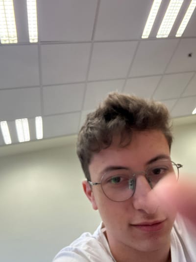 A young man with curly brown hair, wearing round glasses and a white shirt, looks down at the camera. A finger is partially obscuring the view of his face. The background features a ceiling with recessed lighting panels, suggesting an indoor setting like an office or classroom. The angle of the shot is from below.