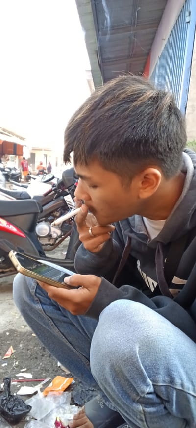 In Kiangroke, Indonesia, a young man with short, dark hair is captured in a close-up shot, crouched down and intensely focused on his smartphone. He is holding a lit cigarette to his lips, with a wisp of smoke curling upwards, suggesting he is in the process of exhaling. His right hand, adorned with a silver ring, is positioned near his mouth, possibly steadying the cigarette or gesturing. The man is dressed in a dark grey hooded sweatshirt over a white t-shirt and light blue jeans. The background reveals a street scene with several motorcycles parked, indicating a common mode of transportation in the area. The environment appears to be outdoors, possibly in a marketplace or a casual gathering spot, with some debris scattered on the ground, including plastic bags and wrappers, hinting at a busy, lived-in urban setting. The overall impression is a candid moment of everyday life, capturing a blend of modern technology and personal habit against the backdrop of Indonesian street culture.