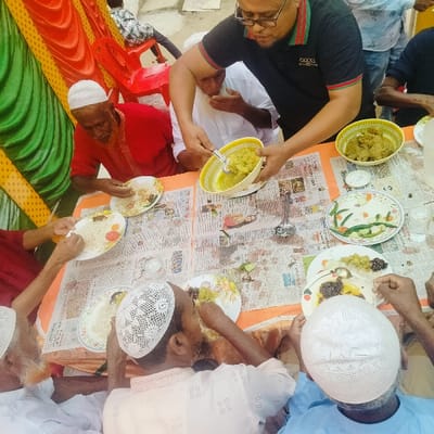 This image depicts a communal meal occurring in Lalmohan, Bangladesh. Eight adult males are observed around a rectangular table covered with newspaper sheets laid over an orange fabric. On the left side, green and orange/yellow drapes suggest a temporary or festive gathering. One man, wearing a black polo shirt with "GUCCI" branding and glasses, stands and serves a light green-yellow curry from a patterned yellow bowl using a metal ladle. Seven other men, dressed in traditional attire including various white prayer caps, are seated around the table. They are actively consuming food—rice and various curries—from individual white plates using their hands. Additional food items on the table include a larger bowl of dark green/brown curry and a white plate containing sliced cucumber, green chilies, and carrots. A glass of water is also visible. Red plastic chairs and a concrete-like surface are present in the background.