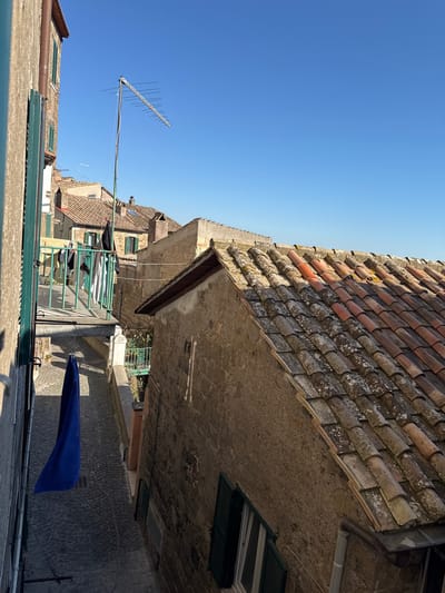The image shows a narrow street in Castelnuovo di Porto, Italy, viewed from a high vantage point, possibly a rooftop or window. The street is lined with old buildings, characterized by tiled roofs, stone walls, and green-shuttered windows. Laundry is hanging on a drying rack between buildings. A blue cloth is draped over a line or structure on the right side of the street. An old television antenna is visible against a bright blue sky. The street surface is paved with cobblestones.