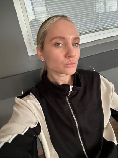 The image displays a Caucasian female subject positioned indoors, located in Kemalpaşa, Turkey. Her head and upper torso are visible, with her gaze directed towards the camera. The subject has light blonde hair, pulled back from her face, revealing a fair complexion, light eyebrows, and green eyes. A silver-colored hoop ring is visible in her right nostril. Her lips are slightly pursed. She is dressed in a long-sleeved, collared jacket with a full-length, silver-toned zipper. The jacket is primarily black, featuring broad cream-colored vertical panels on the sides and sleeves. The background consists of a plain grey wall section. To the subject's upper left, a white-framed window fitted with horizontal blinds is visible. A dark grey horizontal surface, potentially a ledge or counter, extends beneath the window, featuring two small metallic hooks positioned behind the subject's right shoulder.