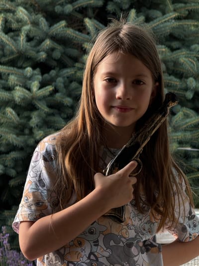 A young girl, with long brown hair, is positioned in the center of the frame, her face tilted upwards. She wears a light-colored shirt adorned with a pattern of cartoon cats in various poses and expressions. Her right arm is raised, holding a toy pistol with a black barrel and gold accents. The pistol is held close to her head, with her fingers wrapped around the handle. Her gaze is directed slightly away from the camera, and she has a neutral expression.

The background is dominated by the dark green, blurred foliage of a coniferous tree, suggesting an outdoor setting. The lighting appears natural, hinting at daytime, possibly late afternoon due to the shadows and highlights on the girl's skin and hair. The overall impression is that of a playful moment, with the girl engaging with the toy. The image is oriented in landscape mode, with the girl's head towards the left of the frame. No visible text is present in the image.