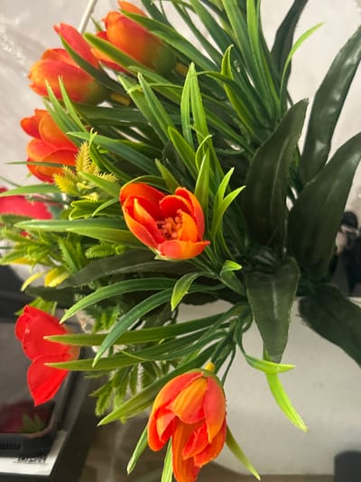 The image shows a close-up of a bouquet of artificial tulips with vibrant orange and red petals and green leaves. The flowers are arranged in a way that suggests they are part of a larger display, possibly on a table or in a vase. The background is blurred and light-colored, with some hints of darker shapes that could be furniture or other objects. There is no indication of the city of Georgiyevskaya, Russia in the image, nor any people or actions.