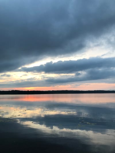 This is a vertical, eye-level, outdoor, medium shot photograph of a tranquil lake at sunset, with a dark tree line in the background. The sky is filled with dramatic, heavy grey clouds that are beginning to break up near the horizon, revealing a soft, warm glow of orange and pink from the setting sun. The lake's surface is calm and reflective, mirroring the colors of the sky and the dark silhouette of the trees, creating a serene and somewhat moody atmosphere. The location is identified as Svirstroy, Russia. There are no people, actions, or interactions depicted in the image. The environment is a natural landscape dominated by the sky and water.
