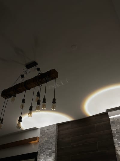 The image displays an interior scene, likely a commercial or residential space, under dim lighting conditions. A prominent feature is a rustic-industrial style chandelier, horizontally suspended from a white ceiling by black chains and hardware. The chandelier consists of a dark wooden beam from which approximately ten or more Edison-style filament bulbs hang at varying lengths via black cords. Most bulbs are illuminated, casting warm light. Circular light reflections with distinct rainbow-like edges are visible on the white ceiling and a partition wall to the right, emanating from light sources not fully visible. The lower right portion of the image reveals a partial view of a wall or partition structure. This structure features a lower section composed of gray brick-like or stacked stone-like elements and an upper section with dark, horizontally oriented rectangular panels, possibly wood or wood-look material. A lighter colored, horizontal wooden beam or shelf is visible protruding from the wall in the lower left background. No individuals are present in the frame. The scene is located in the Arvot Hayarden Regional Council, Palestinian Territory.