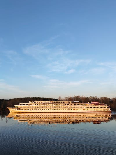 A long, white passenger ship, "Konstantin Korotkov" in Cyrillic script, is shown on a body of water, likely a wide river or lake. The ship has multiple decks of passenger windows, is reflected in the water below, with the opposite shore in the background. The trees lining the shore are mostly bare, suggesting a fall or early spring season. The sky is a clear light blue with wispy clouds, indicating clear weather. The image appears to be taken at sunrise or sunset, evidenced by the warm lighting on the ship and its reflection. The location, 3-Y Uchastok, Russia, is not evident in the image.