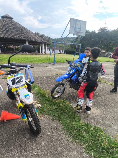 The image depicts an outdoor scene in Timaná, Colombia, on a concrete and grassy area. In the foreground-left, a white and yellow Husqvarna dirt bike with the number "12" and "MARTÍN RIVA" visible on its front plate is parked, with an orange traffic cone resting near its front tire. To the right, a blue Yamaha dirt bike with the number "61" is stationary. Two children are central to the frame; one child, positioned with their back largely to the viewer, wears full motocross gear including a black and white checkered top with a chest protector, red and black pants, and white motocross boots, and faces towards a second child. The second child, dressed in a white long-sleeve jersey, stands next to the blue dirt bike. A third individual, whose lower torso and legs are visible, stands on the far right wearing dark clothing. In the background, a blue metal basketball hoop with a wooden backboard is visible. Behind it, a blue metal soccer goal frame stands. Further back, a large gazebo with a dark, possibly thatched roof supported by white pillars is visible on the left, and white buildings with red roofs are scattered across the mid-ground. The area features concrete ground with interspersed patches of green grass and weeds. Distant green hills and trees are visible under a partly cloudy sky.
