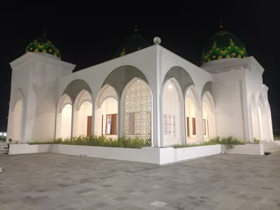 The image shows a mosque at night in Keureuweungkrueng, Indonesia. The mosque is primarily white with green and yellow tiled domes. It features a series of arched entrances with ornate white screens. There are also wooden-shuttered windows visible. A person is seated on steps to the left of the mosque, appearing to be reading or engaged in a quiet activity. The foreground is paved with grey square tiles, and there are low planters with greenery in front of the mosque's facade. The sky is dark, indicating nighttime.