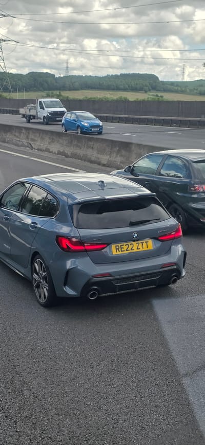 The image shows a grey BMW 1 Series hatchback with the license plate RE22 ZTT, driving on a highway. The car is in the foreground, angled towards the right of the frame. The rear of the car is clearly visible, including its taillights and exhaust pipes. Behind the BMW, a dark-colored car is partially visible. Further in the background, a blue Ford Fiesta and a white flatbed truck are driving on the same highway. The highway appears to be elevated, with a concrete barrier separating lanes and a retaining wall visible. Beyond the highway, there is a wooded area and a cloudy sky. There are also power lines and pylons in the background. The scene suggests a typical highway traffic situation. The image does not provide specific details that would allow for pinpointing the exact location within Rotherham, United Kingdom, but it is consistent with a major road in the area.