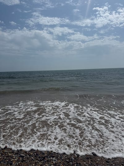 The image captures a close-up view of the sea lapping onto a pebble beach. The scene is divided horizontally. The top half of the image shows a sky filled with a mix of wispy and more dense white clouds against a pale blue background. The bottom half depicts the ocean, with the water appearing dark and slightly rippled. The edge of the water meets the shore, creating a foamy, white surf that washes over a dark, pebbly beach. The pebbles are of various sizes and colors, including grays, browns, and some darker stones. The sun appears to be reflecting off the surface of the water, creating small glints of light. The image is oriented vertically, suggesting it was taken in portrait mode. No people, objects, text, or specific activities are visible. The weather appears to be cloudy but bright, possibly indicating a sunny day with scattered clouds. The location is Youghal, Ireland, which is known for its coastline and beaches. The overall mood is serene and natural, focusing on the elements of sea, sky, and shore.