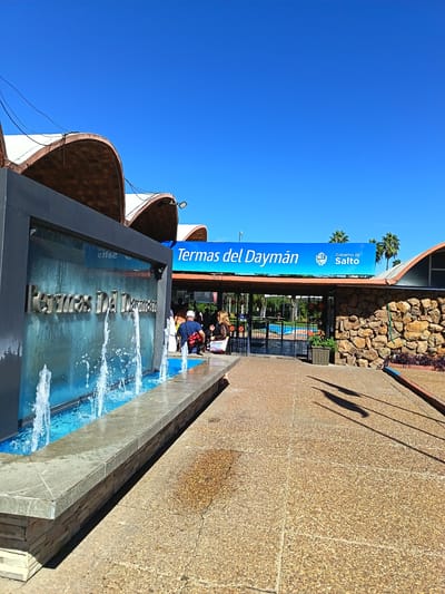 The photograph depicts the entrance to Termas del Daymán, a hot springs resort located in the city of Termas del Daymán, Uruguay. A blue sign reading "Termas del Daymán, Gobierno de Salto" is prominently displayed above the entrance. A fountain with the name Termas Del Dayman is visible on the left side of the entrance. The entrance is a stone archway. People are visible near the entrance, including individuals wearing swimsuits. The sky is clear and blue. The ground is paved with a textured surface. Shadows are cast onto the ground, indicating sunlight. Palm trees are visible in the background. The building is made of stone.