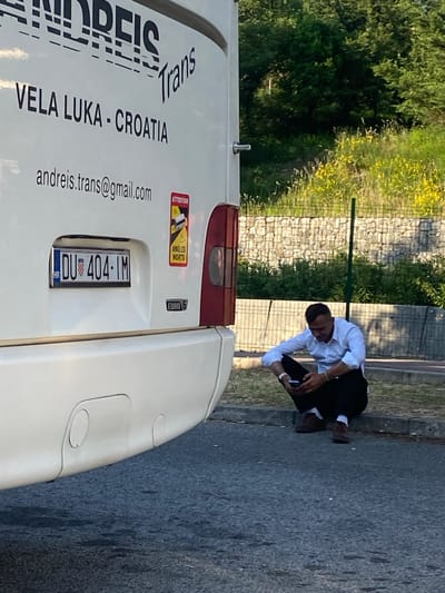 A man is crouching and looking at a smartphone next to a white van on an asphalt road in what appears to be a semi-rural area. The back of the van has "ANDREIS Trans" and "VELA LUKA - CROATIA" printed in bold lettering, along with "andreis.trans@gmail.com" and the license plate "DU 404-IM." There are also warning stickers and a "EURO 6" label on the van's rear. In the background, there is a vegetated area with a retaining wall and trees. Based on the information from the license plate and the text on the van, the location is likely near Vela Luka, Croatia. There is no evidence to suggest that this photo was taken in Palje Brdo, Croatia.