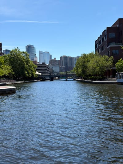 This is a photograph taken on a clear, sunny day, looking down a wide river that flows through a city. The river occupies the majority of the foreground, with its surface reflecting the blue sky and showing small ripples. On the left bank, there is a stone walkway with a rounded edge and some vegetation. On the right bank, there is a brick building with multiple windows and a balcony, as well as a white boat with windows and an American flag visible at the far right of the frame.
Further in the distance, across the river, a bridge spans the water, with traffic visible on it. Beyond the bridge, a cityscape of modern buildings and skyscrapers rises against the bright blue sky. Trees with lush green foliage line the riverbanks, softening the urban landscape. The scene appears to be the Charles River in Cambridge, Massachusetts, specifically a view towards the Boston skyline. There are a few indistinct figures of people walking along the riverfront on the right side.