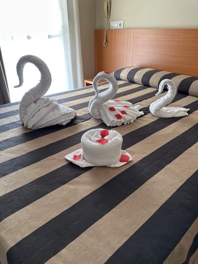 The image shows a bed with four towel sculptures arranged on it. The sculptures are white and appear to be shaped like swans. Two of the swans have red petals decorating their necks and heads. The bed is covered with a striped comforter in shades of beige and dark grey. The scene is indoors, likely in a hotel room in Lloret de Mar, Spain, as indicated by the location context. One hint image shows a wall-mounted telephone and light switches, further suggesting a hotel room setting. The lighting in the image is bright, hinting at daytime. There is no visible text in the image. The overall impression is one of a welcoming and decorative touch for guests.