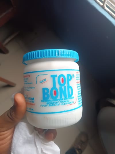 A close-up, eye-level shot captures a person's hand holding a jar of "TOP BOND" general-purpose white glue in an indoor setting in Warri South, Nigeria. The white, squat plastic jar features a bright blue, ribbed screw-on lid. The label prominently displays the brand name "TOP BOND" in large blue letters with red "O"s, accompanied by a small "NEW" indicator. Below the brand name, "GENERAL PURPOSE WHITE GLUE" and its French translation, "NOUVELLE FORMULE COLLE BLANCHE MULTI-USAGES," are printed.

On the left side of the label, partial text under a "SAFETY" heading is visible, including words like "use," "equipment," "children," and "skin." Below this, fragments of a company name, "ECHEM LIMITED," and "NIGERIA" are discernible. On the right side, headings like "USES" and "HOW TO" are present, with partially visible instructions and applications such as "Good for" and "application."

The hand holding the jar is light-skinned, with a white, tissue-like material partially visible beneath it. The background is dimly lit, suggesting an indoor environment, possibly a room, with dark shadows and hints of brighter light filtering in from an unseen window or opening on the right side. Part of a person's foot, wearing dark footwear, is visible on the floor in the lower right corner, suggesting they are seated or standing. The time of day appears to be daytime due to the natural light.