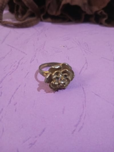 A silver-toned metallic ring, designed with a multi-petaled floral motif, rests on a flat, light purple surface that exhibits faint, irregular linear textures. Several small, clear, possibly stone elements are visible at the center of the floral design. A distinct shadow is cast to the left of the ring, indicating a light source from the upper right. The background features an out-of-focus, dark brown, irregularly shaped textile material, likely fabric. The scene is located in the city of Chandragiri, Nepal.