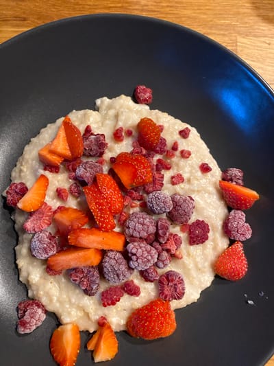 A top-down, close-up shot depicts a black plate filled with what appears to be porridge, topped with cut strawberries and frozen raspberries. The plate rests on a wooden surface. The image lacks any information indicating its location in Lommedalen, Norway, or any presence of people, actions, or interactions.