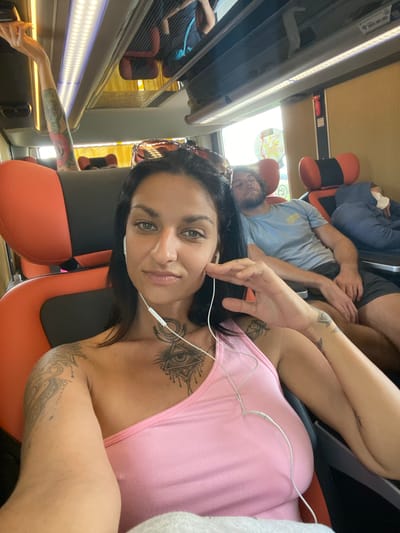 In the city of Ninh Hoa, Vietnam, a woman with dark hair and extensive tattoos takes a selfie on a bus. She wears a pink one-shoulder top and white earbuds. To her right, a man with light brown hair sleeps in his seat, wearing a blue t-shirt and dark shorts. Further back, another person is asleep, covered by a blue hoodie. The bus interior features orange and black seats and bright overhead lighting. A window on the right side of the bus shows a blurred, sunny outdoor scene.