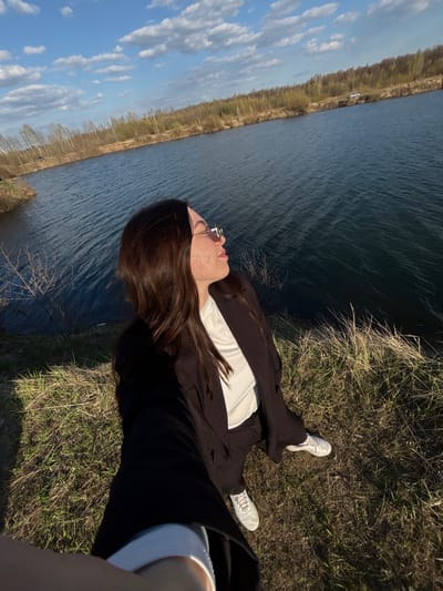 A woman lies on her back on a grassy bank next to a body of water under a blue, partly cloudy sky. The water extends to the left edge of the frame, with a line of trees visible on the far side. The woman is wearing white sneakers, black pants, a white shirt, and a black jacket. She has long brown hair and is wearing sunglasses. The photograph appears to be taken in Zaosinovo, Russia, based on visual details and the presence of a body of water adjacent to a forested area, which is typical of the region.