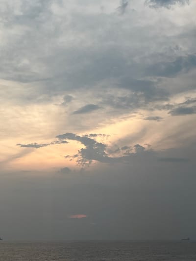 The image depicts a cloudy sky over a body of water. The clouds are varied in texture and color, ranging from light gray to darker shades, with patches of orange and yellow light breaking through in the middle section. The water surface is rippled. Two ships are visible in the distance, one on the left and another on the right, appearing as small silhouettes against the horizon. The scene has a muted color palette, suggesting it might be during dawn or dusk. The location is identified as Unknown, Malaysia.
