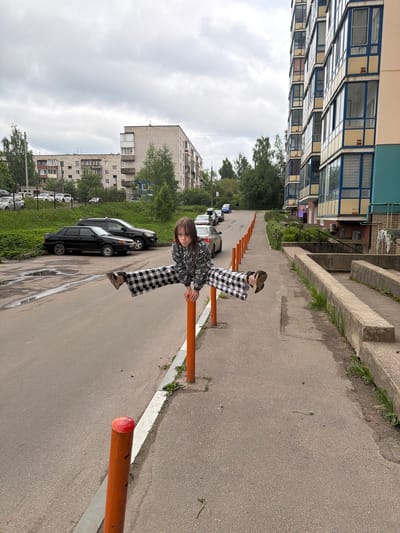 A child sits on an orange barrier along an asphalt road in Tokkari-Lend, Russia. The child wears black and white checkered pants and a grey patterned jacket. A row of orange barriers lines the road. Several cars are parked along the road behind the child. Buildings are visible in the background. The sky is overcast.