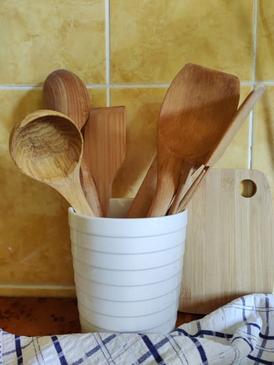 In a domestic kitchen setting, likely in Rybnitsa, Moldova, a white ceramic utensil holder stands on a patterned countertop. The holder contains several wooden kitchen utensils, including spoons and spatulas, exhibiting natural wood grain. To the right of the holder, a light-colored bamboo cutting board with a circular cutout rests against the tiled wall. The wall is covered in glossy, yellow square tiles. A blue and white checkered cloth is draped in the foreground. The lighting suggests an indoor environment, possibly natural light from a window.