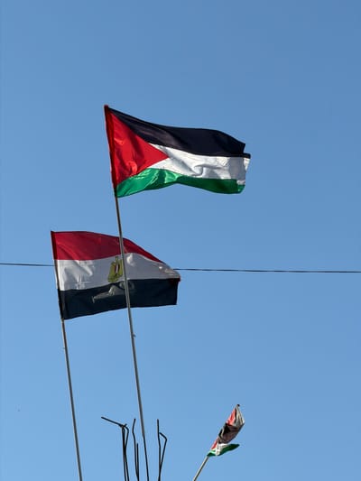 In Jabalia, Palestinian Territories, against a clear blue sky, three flags are visible. The uppermost flag is the Palestinian flag, with horizontal stripes of black, white, and green, and a red triangle on the hoist side. Below it, to the left, flies the Egyptian flag, featuring three horizontal stripes of red, white, and black, with the Eagle of Saladin centered on the white stripe. To the far right and lower, a small section of the Palestinian flag is also visible on a pole. Two tall poles, one supporting the Palestinian flag and another behind the Egyptian flag, are present. Additional thin poles and dark, wire-like objects are clustered near the base of the flags, suggesting a makeshift or temporary setup. A dark horizontal line, likely a wire, stretches across the mid-ground of the image.