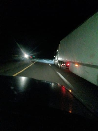 A dark highway at night is illuminated by headlights and the running lights of a large truck traveling in the adjacent lane. White and yellow lane markings guide traffic. The dark hood of a vehicle is visible in the foreground, reflecting the lights on the road. Distant headlights approach in the opposite direction.