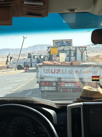 The image, taken from inside a vehicle, shows the view through the windshield, looking towards the arid landscape. The scene features several trucks, including an ISUZU truck with license plate "BRB QD 154," and an Egyptian flag visible on the truck's rear. The license plate and the speedometer are visible in the lower portion of the frame. The OCR extracted text further provides data such as a vehicle performance indication. The background includes a mountain range. This is most likely on the road between Gharib Cape, Egypt and the mountains.