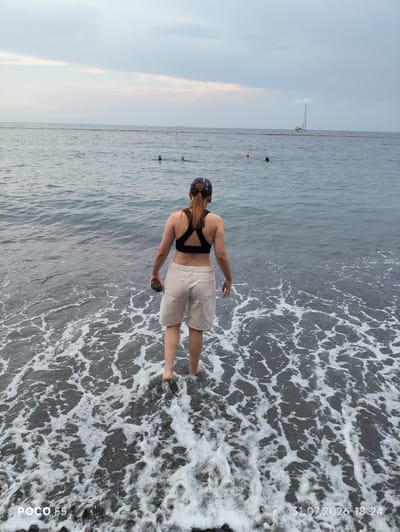 A person stands at the edge of the sea, their back to the camera, with waves lapping at their feet. They are wearing a black sports bra and light-colored shorts, and a baseball cap is on their head. They are holding a small, dark object in their right hand. The sea is choppy, with white foam from the waves creating a textured pattern on the dark sand. In the distance, several people are swimming in the water, and a sailboat is visible on the horizon. The sky is overcast with hints of pink near the horizon. The timestamp "31.07.2026 18:24" and "POCO F5" are present in the lower corners of the image.