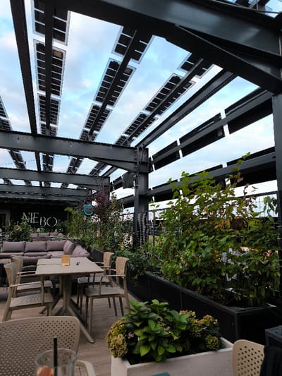This image depicts a modern rooftop terrace, specifically the Niebo bar/restaurant located at ul. Wrocławska 1A, Oława, Poland.
The scene is an outdoor seating area under a pergola structure. Overhead, a black steel framework supports numerous transparent solar panels arranged in parallel rows, allowing a view of a partially cloudy blue sky.
The terrace features two primary seating arrangements:
1. In the background, a lounge area consists of multiple modular gray sofas. A large black banner with the stylized white text "NIEBO" is prominently displayed behind this area, with a blue spherical light fixture hanging nearby.
2. In the mid-ground, a dining section comprises several light-colored rectangular tables, each surrounded by matching light-colored chairs. One table displays a small gold-colored object.
Vegetation is integrated throughout the space. Large green shrubs, some showing yellowing leaves, are contained within black rectangular planters that line a perimeter, partially obscured by a black metal grid fence. In the immediate foreground, a white planter holds green hydrangea plants with pale pinkish-green blooms.
A partial view of a glass with a straw and a citrus slice, suggesting a beverage, is visible on a light-colored surface in the extreme foreground. The floor of the terrace appears to be light-colored decking. No individuals are present or engaged in any activity within the frame.