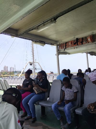 The image captures a scene aboard a passenger ferry or large boat, with coastal urban development visible in the background, consistent with Mkomani ward, Kenya. The perspective is from within the vessel's covered passenger area, looking towards the port side and forward.
The ceiling is a light grey surface, featuring exposed fluorescent tube lighting fixtures and electrical conduits running along its length. A white net containing multiple orange life jackets is suspended from the ceiling structure on the right side. Rows of molded grey plastic seating are arranged on a dark green floor.
In the mid-ground, from left to right, three primary subjects are seated. An adult female with dark hair styled in an updo, wearing a black t-shirt and pink trousers, is holding and observing a smartphone. Adjacent to her, a second adult female with long dark braided hair, wearing sunglasses on her head, a black t-shirt, blue jeans, and white earphones, is also focused on a smartphone in her hands, with a small striped bag resting on her lap. To their right, a young boy with short dark hair, dressed in a white and blue striped t-shirt and blue jeans, sits looking down at his hands. He wears grey slides.
Further back, additional passengers, primarily adults, occupy other seats, with some individuals partially obscured. One individual on the far right wears a white patterned cap. Outside the vessel, a bright blue sky is visible above calm water. The upper structure of another vessel, including a mast and rigging, is prominent. On the distant coastline, a cluster of multi-story buildings indicates an urban waterfront.