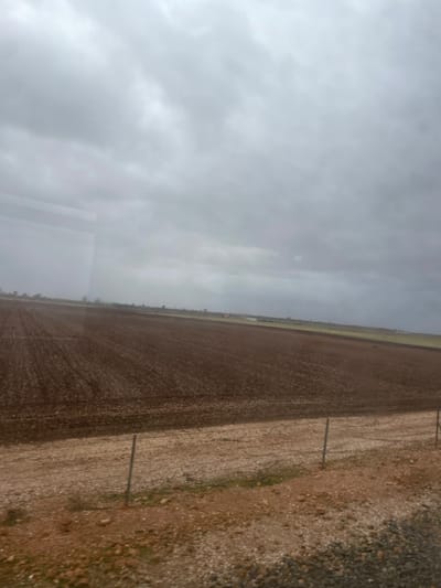 A landscape under a cloudy sky shows a vast, tilled field with parallel furrows stretching into the distance. The soil is a dark brown color. To the right of the field, there is a strip of rough, gravelly ground with sparse patches of green vegetation. Metal posts are sparsely placed along the boundary of the field. The perspective suggests the photo was taken from an elevated position, possibly through a window.