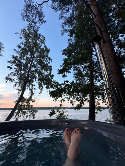 A low-angle, close-up shot shows a person's feet submerged in the dark, rippling water of an outdoor hot tub. The feet are illuminated, with toenails painted bright red. The hot tub's dark gray rim is visible in the foreground. Beyond the tub, a wide body of water stretches towards a tree-lined horizon. The sky above is a gradient of pale blue, transitioning to soft pink and orange hues near the horizon, suggesting dusk or dawn. Tall trees with dark green foliage frame the scene, with a metal chimney pipe positioned to the right, angled upwards alongside a tree trunk. The environment appears to be natural, likely a lakeside setting in Priedesmuiža, Latvia, characterized by trees and water.