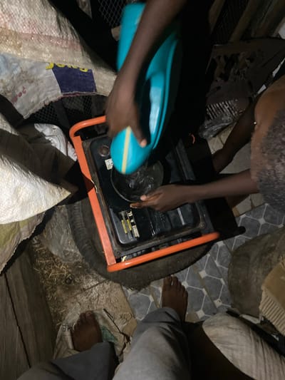 This image provides an overhead perspective of a dimly lit utility area within the city of Igarra, Nigeria, depicting an act of refueling a portable generator.

At least three individuals are partially visible. In the immediate foreground, a person's bare feet, clad in light grey trousers, are positioned on a dirty white woven sack situated on a mixture of earth and straw-like debris. To the right, two dark-skinned individuals are actively engaged with the generator. One person uses their right hand to hold a turquoise container, distinguished by a yellow stripe, from which a liquid is being poured into the generator's fuel tank. Another hand, belonging to a second individual, is visible on the left side of the generator, appearing to steady the unit or guide the pouring process. A dark-skinned arm is also visible in the far right background.

The central subject is a portable generator, characterized by an orange tubular frame and a black engine casing, with a visible "6L" label indicating its capacity. The generator rests on a black rubber tire. The ground directly beneath the generator and extending to the right features a patterned grey and white tiled surface. To the left and under the foreground person's feet, the ground is an uneven mix of dirt, organic debris, and multiple dirty white woven sacks. One sack displays a faded blue and yellow graphic along with red markings. The background is predominantly dark, revealing some metal mesh grating in the upper left.