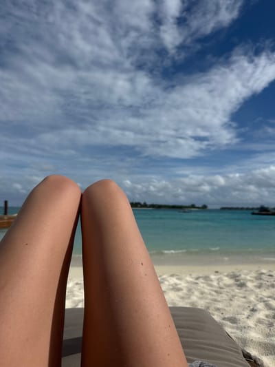 The image is a first-person perspective of a person relaxing on a beach lounge chair. The foreground is dominated by the person's tanned legs, with visible skin texture and a few moles. In the mid-ground, a pristine white sandy beach meets the turquoise ocean. The water is calm, with small waves lapping at the shore. In the background, a thin strip of land with palm trees and some buildings is visible, suggesting a resort or island setting. The sky is a vibrant blue, adorned with scattered white clouds, typical of a sunny day. The overall scene evokes a sense of tropical relaxation and vacation. The location is identified as Unknown, Maldives, a generic tropical island destination.