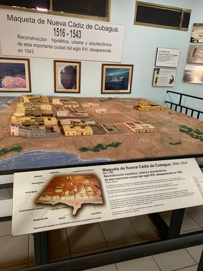 The image displays a museum exhibit featuring a detailed architectural model of Nueva Cádiz de Cubagua, a city that existed from 1516 to 1543. A large text panel above the model provides its title, "Maqueta de Nueva Cádiz de Cubagua. 1516-1543," and a description stating it is a "Reconstrucción hipotética, urbana y arquitectónica de esta importante ciudad del siglo XVI, desaparecida en 1543." Below the model is a diagram with labels identifying key areas of the city, such as "MERCADO," "CEMENTERIO," "CONVENTO DE SAN FRANCISCO," "AYUNTAMIENTO," "PLAZA MAYOR," "IGLESIA MAYOR DE SANTIAGO," "CALLE REAL LA MARINA," "BATERIA MILITAR," "MUELLE," and "CASA FUERTE." Several framed photographs and smaller informational plaques are mounted on the wall behind the model, offering visual and textual context about the city's history and ruins. The environment appears to be an indoor exhibition space, with the model situated on a table with a dark railing in the foreground. The lighting suggests an indoor setting with artificial illumination. The provided information does not indicate the specific location of Boca del Río, Venezuela, as being associated with this exhibit; rather, the exhibit itself pertains to Nueva Cádiz de Cubagua.