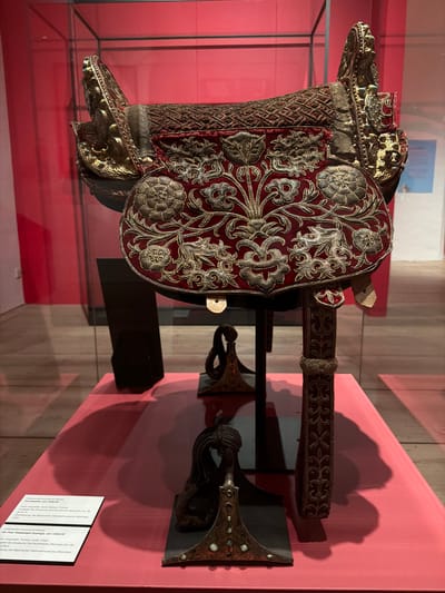 The image captures an elaborately decorated saddle and a pair of matching stirrups displayed in a museum or exhibition setting. The central object is a "Promksattel" (saddle), dated around 1600/20 according to the accompanying text. It is crafted from wood, silver-plated components, and rich red velvet, which is extensively embroidered with intricate gold and silver threads forming floral and foliate patterns, including distinctive rose motifs. The front pommel and rear cantle feature ornate gilded metalwork, possibly depicting mythological creatures or dragons, accented with small turquoise or ruby-like gemstones. A decorated strap or stirrup leather is also visible.
Adjacent to the saddle are "Ein Paar Steigbügel (Üzengi)" (a pair of stirrups), also dated around 1600/20. These stirrups are described as gilded, and composed of turquoise, leather, and silver, displaying a similar level of luxurious detail and craftsmanship.
The artifacts are presented on a red-covered pedestal against a plain red wall, likely within a glass display case, as indicated by reflections. Shadows are cast on the wall behind the items. The visible text plaques confirm that both the saddle and stirrups are loans from the Museum Fünf Kontinente, Munich, and transferred from the Bayerisches Nationalmuseum, Munich, underscoring their historical and artistic value. The display highlights the exquisite artistry and opulence associated with equestrian gear of that period, likely belonging to an individual of high status.