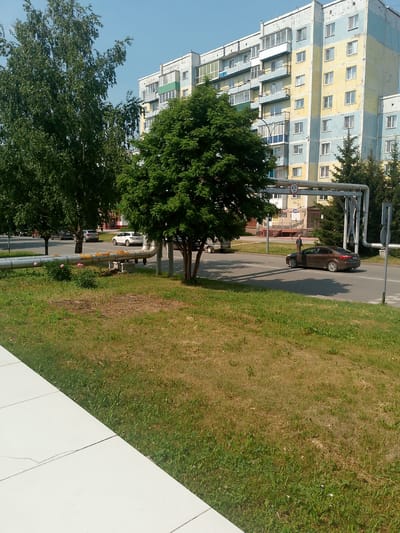 The image shows a street scene in Polysayevo, Russia. In the background, there is a multi-story apartment building with a yellow and light blue facade. In the foreground, a brown sedan is turning onto a street, with a pedestrian standing near the building entrance. A large pipe runs horizontally across the middle of the image, supported by metal structures. Trees and a grassy area are also visible.