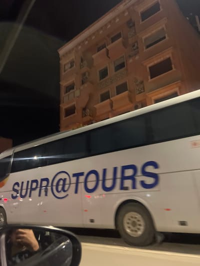 The scene depicts an urban environment at night in Beni Mellal, Morocco. A white passenger bus, branded "SUPR@TOURS" in blue capital letters, occupies the central and lower portions of the frame, moving from left to right. The bus features tinted windows and is partially blurred, indicating motion. In the background, a multi-story building, appearing light orange or peach in color, rises above the bus. This building has multiple windows, some with decorative sills or surrounds, and several small, ornate balconies or architectural projections. Multiple external air conditioning units are affixed to its facade. The image is captured from inside another vehicle, with the upper edge of a car door frame visible across the lower part of the building and the top of the bus. The vehicle's left side mirror is visible in the bottom-left corner, reflecting a hand, presumably of the photographer. The overall illumination is artificial, suggesting streetlights or building lights against a dark night sky.