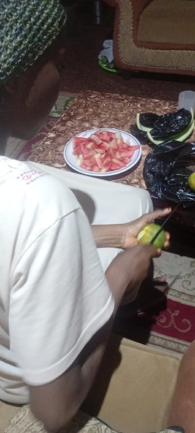 A person is seated on the floor in a room located in Rijiyar Zaki, Nigeria, wearing a white shirt and a patterned cap. The person is peeling a green fruit with a knife. On the floor in front of them is a white plate containing diced watermelon. To the right of the plate is a black plastic bag and cut watermelon rinds on a green plate. A patterned rug is visible on the floor. A sofa partially visible in the background.