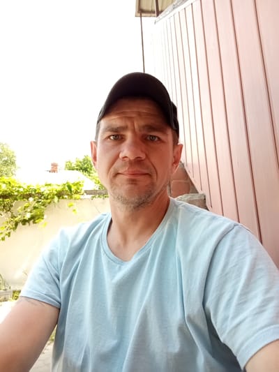 The image is a selfie taken outdoors in Solonytsivka, Ukraine. A man wearing a light blue t-shirt and a black baseball cap is in the foreground, facing the camera. He has light skin, light-colored eyes, and stubble on his chin. In the background, there is a light-colored wall, a vine with green leaves, a portion of a building with a red-tiled roof, and a light pink, vertically paneled structure. The sky is bright and overcast. The man's left arm is visible, showing a tattoo on his forearm.