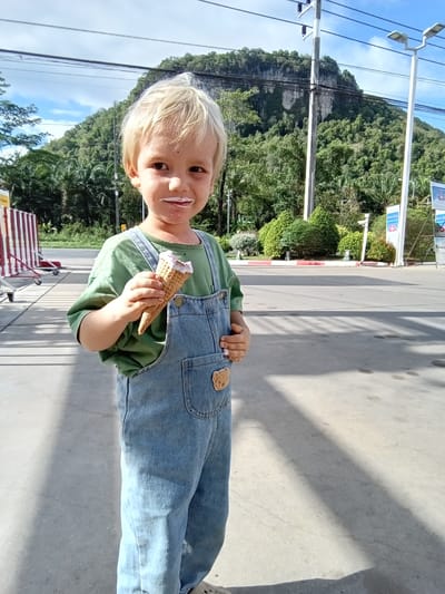 A young boy with blonde hair, wearing a green t-shirt and denim overalls, stands outdoors holding an ice cream cone with a bite taken out of it. He has ice cream on his face. In the background, a lush green mountain covered in trees dominates the scene. There are power lines and streetlights visible, along with a road and some signs, suggesting a public area. The ground is paved concrete, and shadows from the overhead structures are cast across it. The location is identified as Tambon Tha Uthae, Thailand.