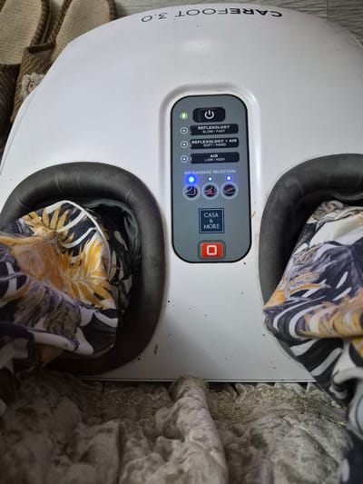 In the city of Qanaq, Kazakhstan, a white foot massager with the brand name "CAREFOOT 3.0" is positioned on a soft, textured floor covering. The massager features a control panel with several illuminated buttons and indicators. One button, marked with a power symbol, is illuminated green. Below this are three selectable modes: "REFLEXOLOGY SLOW/FAST," "REFLEXOLOGY + AIR SOFT/HARD," and "AIR LOW/HIGH." The "AIR MASSAGE SELECTION" section displays three illuminated blue squares, suggesting that a selection has been made. The massager has two openings, each lined with a padded cushion, where floral-patterned fabric is inserted. The fabric exhibits a pattern of large, stylized leaves in shades of yellow, orange, black, and white. The overall scene suggests a domestic setting, possibly a living room or bedroom, given the presence of what appear to be slippers in the background and the soft floor covering. The image is taken from a slightly elevated, top-down perspective.