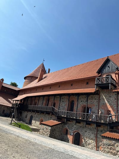 The image depicts the Trakai Island Castle in Trakai, Lithuania. The castle is constructed primarily of red brick and stone, with distinctive terracotta tiled roofs. A wooden walkway with intricate railing details runs along the upper levels of the castle, connecting various sections. Several people are visible on the walkway, some walking and others conversing. In the background, a conical tower with a flag at its peak rises above the main structures. The sky above is a clear, bright blue with two birds in flight. The ground in the foreground is a mix of gravel and paved pathways. The overall scene suggests a sunny day, likely during tourist season, with visitors exploring the historical landmark.