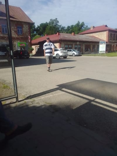 The image shows a man standing on a paved area in front of stone buildings with red tiled roofs. He is wearing a striped polo shirt and shorts. There are cars parked in the background, and trees are visible behind the buildings. A sign with text and a map is posted to the right of the buildings. The location is identified as Koknese, Latvia. A shadow from an overhead structure is cast across the paved area in the foreground.