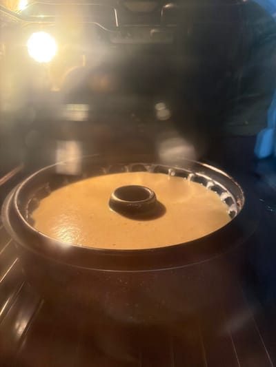 A bundt cake batter is baking in a dark bundt pan inside an oven. The batter is a light yellow color and appears smooth. The bundt pan has a ring in the center and is sitting on a rack within the oven. A bright light from the oven's interior illuminates the scene, creating some glare and reflections on the glass of the oven door. The background is dark and out of focus, showing the interior of the oven and possibly the exterior of other appliances or kitchen elements. The image is taken from inside the oven, looking towards the cake. There are no people or visible signs of the city of Zmeitsa, Bulgaria.